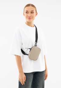 Borsa a tracolla grigia con forma rettangolare, dotata di una robusta tracolla nera e dettagli con zip, indossata sopra una maglietta bianca larga e jeans scuri.