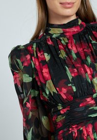 Zwarte bloemenjurk met rode en groene bloemen, met een hoge hals, geplooid ontwerp en doorschijnende lange mouwen. Textuurstof versterkt het detail.