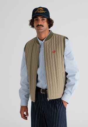 Man with mustache wearing navy cap, beige quilted vest over light blue shirt, and black pinstripe pants, standing with one hand in pocket.
