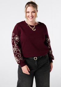 Bordeaux sweatshirt met ronde hals, voorzien van decoratieve crèmekleurige bloemenborduursels op de lange mouwen. Draagt met zwarte jeans en een riem.