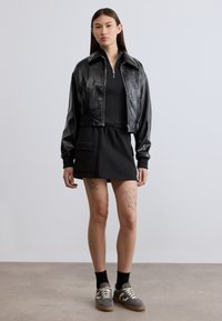 Μαύρο δερμάτινο κοντό μπουφάν, εφαρμοστό μαύρο φόρεμα με τσέπη μπροστά, γκρι αθλητικά παπούτσια και μαύρες κάλτσες. Λεία υφή, casual σχεδιασμός.