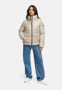 Beige pufferjack met een hoge kraag, een rits aan de voorkant en twee grote voorzakken, gecombineerd met een wijdvallende blauwe spijkerbroek en witte sneakers.