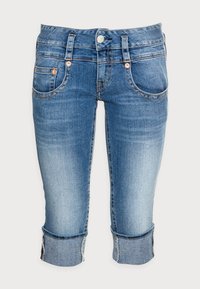 Džínové capri kalhoty světle modré barvy s přiléhavým střihem, pěti kapsami a ohrnutými manžetami. K dispozici jsou s kovovými prvky v zlaté barvě a kontrastním prošitím.