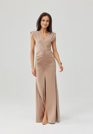 Kobieta z długimi ciemnymi włosami, ubrana w dopasowaną beżową suknię z marszczeniami, z głębokim dekoltem w kształcie litery V i rozcięciem z przodu, stojąca na tle jednolitego jasnego tła.