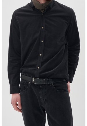Mand iført en sort langærmet knap-op skjorte stoppet ned i sorte jeans med et sort læderbælte og et brunt tørklæde om halsen.