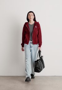 Bordo zip-up dukserica, siva majica, široke svijetloplave traperice i crna torba. Model hoda na neutralnoj pozadini, minimalistički ambijent.