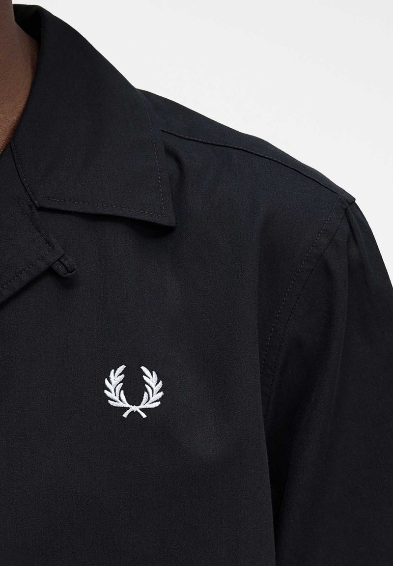 Gros plan sur une chemise noire à col avec un petit logo de couronne de laurier blanche brodé sur la poitrine.