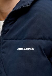 Tmavě modrá prošívaná bunda s bílým logem „JACK & JONES“ na hrudi, se zipovým zapínáním a hladkou texturou.