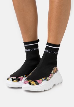 Nohy v černých teniskách ve stylu ponožek s bílými podrážkami a barevnými květinovými vzory na špičce a bocích.
