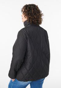 Čierna prešívaná páperová bunda s vysokým golierom, s vlnitým vzorom a voľným strihom. Nosená na svetlomodrých džínsoch.