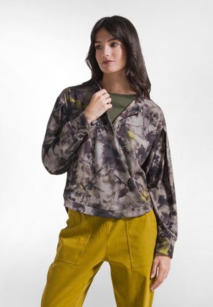 Mujer con cabello oscuro que lleva una blusa de manga larga floral gris y marrón y pantalones amarillos, mirando ligeramente hacia un lado con una mano cerca del cuello.