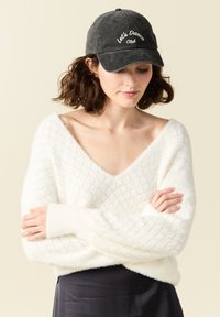 Pullover bianco e soffice con scollo a V e motivo a rombi, abbinato a un cappello nero con testo bianco ricamato "Let's Dance Club."