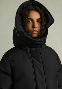 Čierna prešívaná bunda s vysokou, oversized kapucňou a predným zipsovým uzáverom. Obsahuje hladkú textúrovanú látku a detail s knoflíkovým golierom.