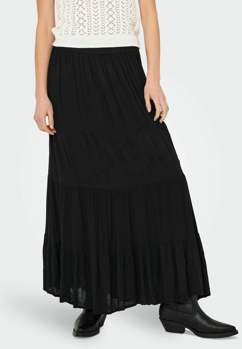 Jupe maxi noire à volants en tissu léger, dotée de détails en œillets sur les coutures et d'un design fluide, associée à des bottes noires à chevilles.