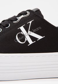 Sneakers in tela nera con suola in gomma bianca. Le caratteristiche includono lacci neri, occhielli in argento e grande logo bianco "CK" sul lato.