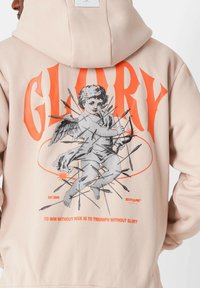 Sudadera rosa claro con un gráfico de un querubín rodeado de flechas, con "GLORY" en letras naranjas negritas arriba. El texto en la parte inferior dice "GANAR SIN RIESGO ES TRIUNFAR SIN GLORIA."