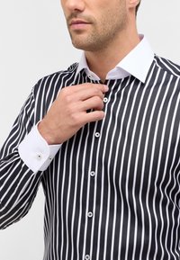 Camisetta a righe bianco e nero con colletto e polsini bianchi. Chiusura frontale con bottoni visibili in argento, design aderente, texture liscia.