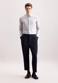 Lichtgrijs overhemd met knopen en kraag, gecombineerd met marineblauwe geplooide broek. De outfit wordt aangevuld met zwarte nette schoenen.