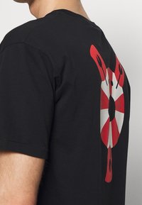 Černé tričko s krátkým rukávem, na zadní straně s červeným a bílým kruhovým vzorem s černými akcenty. Měkký bavlněný materiál.