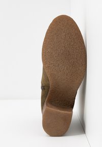 Semelle unique de chaussure marron avec grip texturé et petit talon, positionnée à la verticale contre un mur d’angle blanc et le sol.