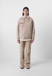 Veste en coton beige avec un col boutonné, associée à un pantalon beige clair et des chaussures en daim. Matériau texturé, coupe ample, design minimal.