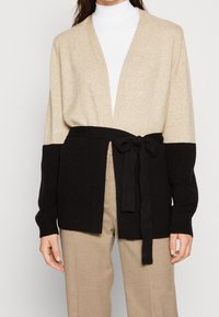 Kvinna som bär en beige och svart färgblockerad cardigan med ett svart bälte över en vit polotröja och beige byxor.