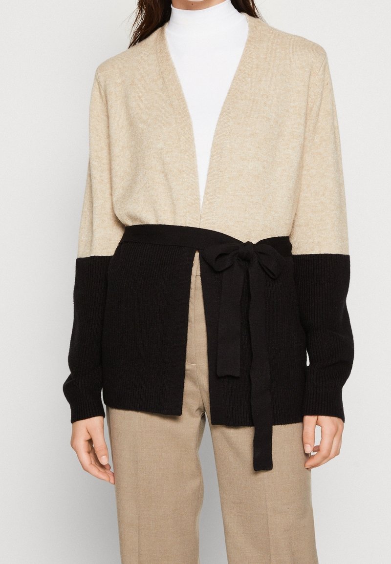 Kvinna som bär en beige och svart färgblockerad cardigan med ett svart bälte över en vit polotröja och beige byxor.