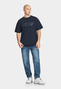 Granatowa bawełniana koszulka z dużym tłoczonym logo "PROSTO", zestawiona z jasnoniebieskimi dżinsami i białymi trampkami z kolorowymi akcentami.