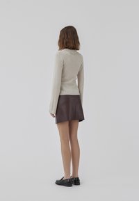 Beige ribbad tröja med hög krage, i kombination med en mörkbrun läder mini kjol. Modellen bär svarta loafers och ses från baksidan.