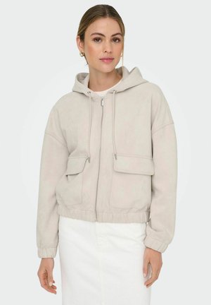 Reißverschlussjacke aus weichem beigem Stoff, ausgestattet mit einer Kapuze, zwei Fronttaschen, elastischen Bündchen und einem cropped Design. Große, glatte Textur, minimale Details.