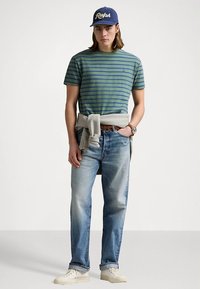 Πράσινο και ναυτικό ριγέ T-shirt, ανοιχτό μπλε τζιν, γκρι φούτερ δεμένο στη μέση, λευκά αθλητικά παπούτσια, ναυτικό καπέλο μπέιζμπολ με λογότυπο.