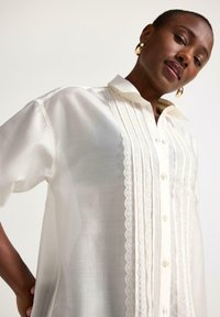 Hvid skjorte i let, gennemsigtigt stof. Har knaplukning foran, blondekant, korte ærmer og klassisk krave.