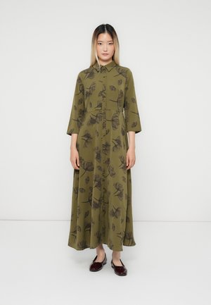 Woman standing against white background wearing olive green, floral-patterned, button-down midi dress and dark red patent flats.