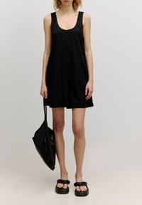 Schwarzes ärmelloses Kleid mit lockerer Passform und Rundhalsausschnitt; getragen mit flachen schwarzen Sandalen und einer großen, glatten schwarzen Tragetasche.