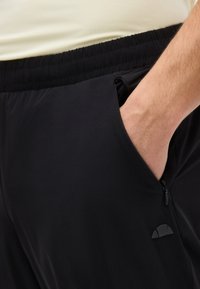 Χέρι στην τσέπη μαύρου παντελονιού με ελαστική μέση και φερμουάρ, με μικρό ημικυκλικό λογότυπο στο πλάι, απέναντι από μπεζ τοπ.