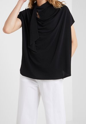 Woman wearing a black draped sleeveless top with a neck tie detail and white wide-leg pants, standing against a plain background.