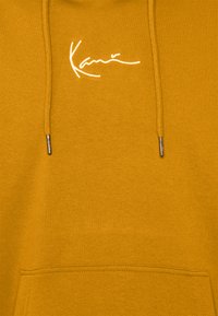 Hoodie v senčeno gorčični barvi, ki ima sprednji žep, vrvico s kovinskimi konicami in belo vezeno logo čez prsni del.