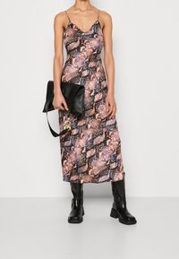 Femme portant une robe nuisette à motif serpent, des bottes noires mi-mollet, et tenant une grande pochette en cuir noir avec un détail en anneau doré.