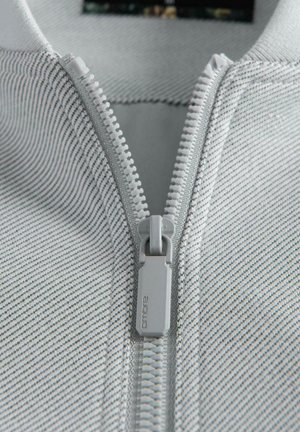 Acercamiento de un zipper de plástico blanco sobre una tela de color gris claro con finas rayas negras, mostrando en detalle el deslizador y los dientes del zipper.