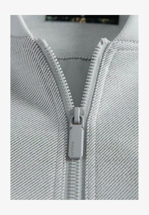 Primo piano di una zip in plastica bianca su un tessuto grigio chiaro con sottili strisce nere, che mostra in dettaglio il cursore e i denti della zip.