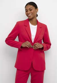 Blazer rosa brillante con revers a scossa, maniche lunghe e due tasche frontali. Abbinato a una t-shirt bianca, con chiusura a un bottone.