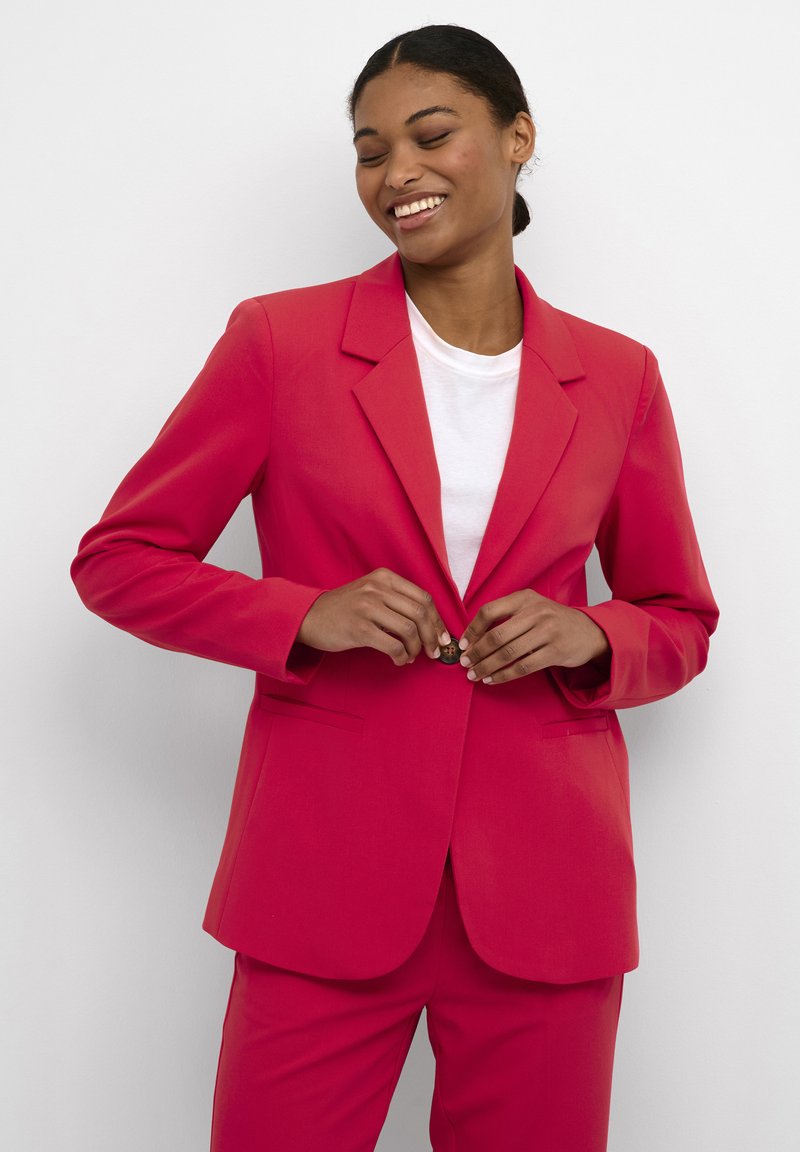 Blazer rosa brillante con revers a scossa, maniche lunghe e due tasche frontali. Abbinato a una t-shirt bianca, con chiusura a un bottone.