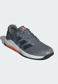 Zapatilla deportiva gris con parte superior de malla, rayas negras y acentos naranjas. Presenta una suela de goma y un collar acolchado para mayor comodidad.