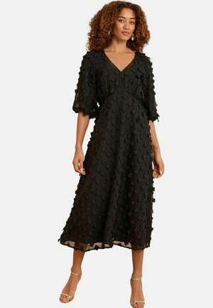 Woman wearing a black midi dress with textured floral appliqués, V-neck, puff sleeves, and gold ankle-strap high heels, standing against white background.
