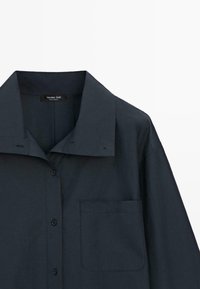 Granatowa, oversize koszula z szerokim kołnierzem, guzikami z przodu oraz kieszenią na piersi po lewej stronie, przedstawiona na białym tle.