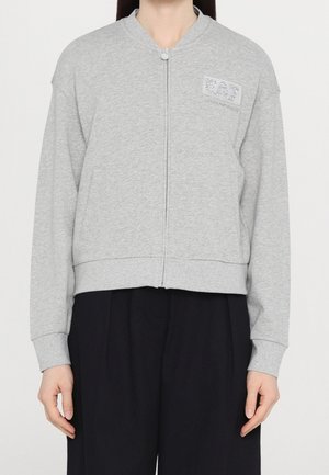 Personne portant un sweat zippé gris clair à manches longues avec des poches, associé à un pantalon noir ample.