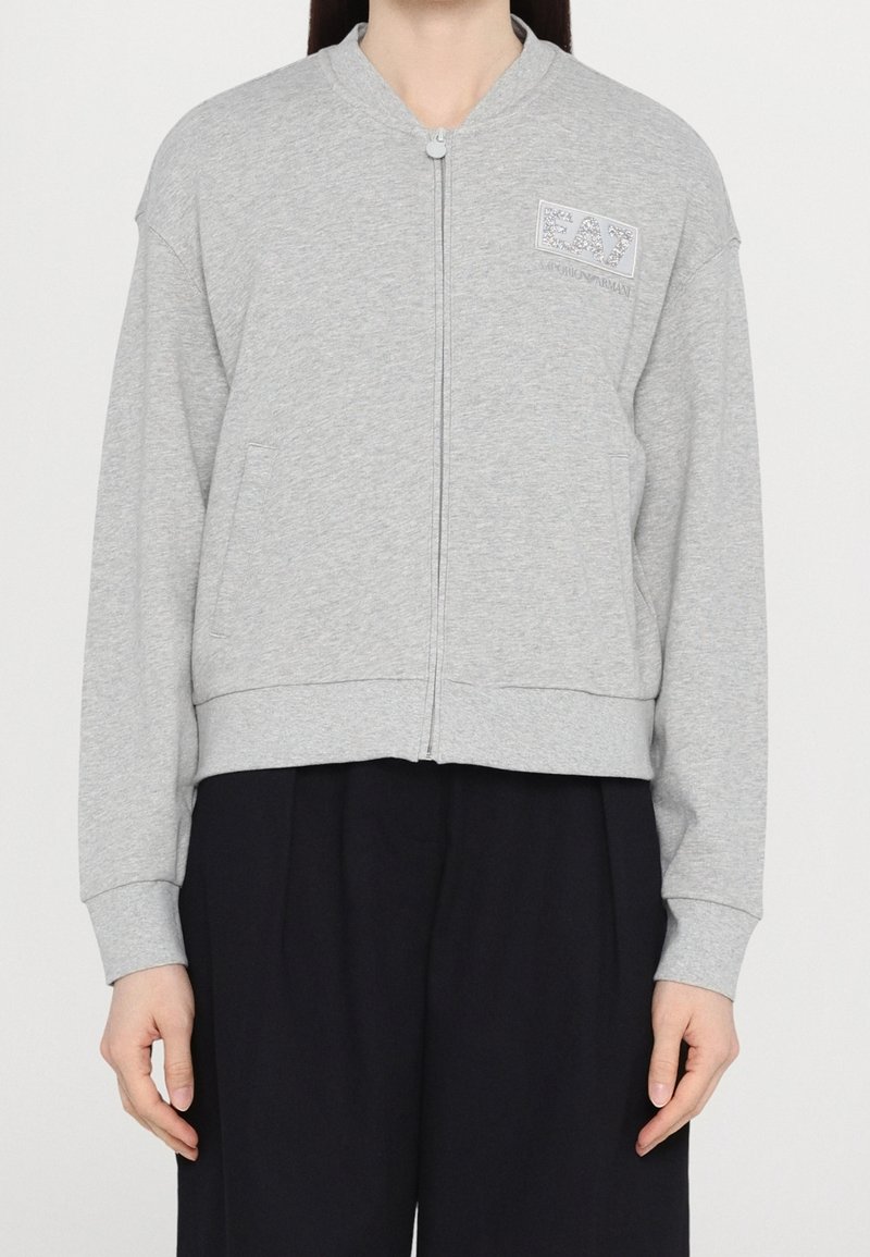 Personne portant un sweat zippé gris clair à manches longues avec des poches, associé à un pantalon noir ample.