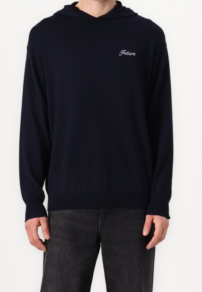 Personne portant un sweat à capuche noir uni avec le mot Future brodé sur la poitrine gauche, associé à un jean noir.