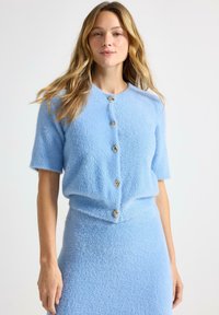 Cardigan azzurro chiaro a maniche corte in tessuto morbido e peloso con sei bottoni color oro sulla parte anteriore. Gonna abbinata visibile sotto.