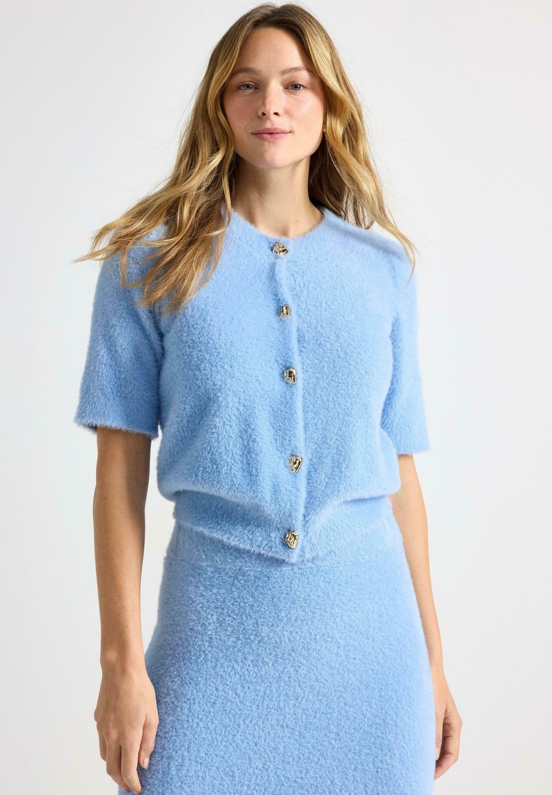 Cardigan azzurro chiaro a maniche corte in tessuto morbido e peloso con sei bottoni color oro sulla parte anteriore. Gonna abbinata visibile sotto.